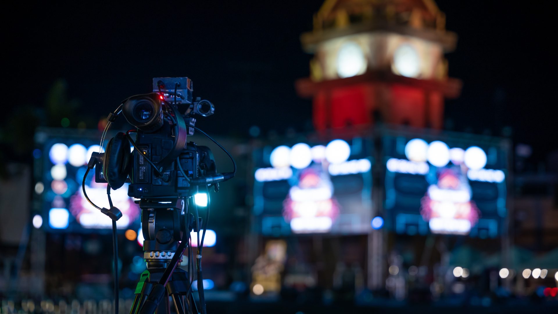 Video production team covers live event on stage. Professional crew utilizes multimedia for movie-like streaming. Expertise in cinematography and live broadcasting, Video equipment "Professional video camera on a tripod at a nighttime corporate event, equipped with a microphone and headphones. Background features illuminated screens and a brightly lit clock tower with an ornate roof."
